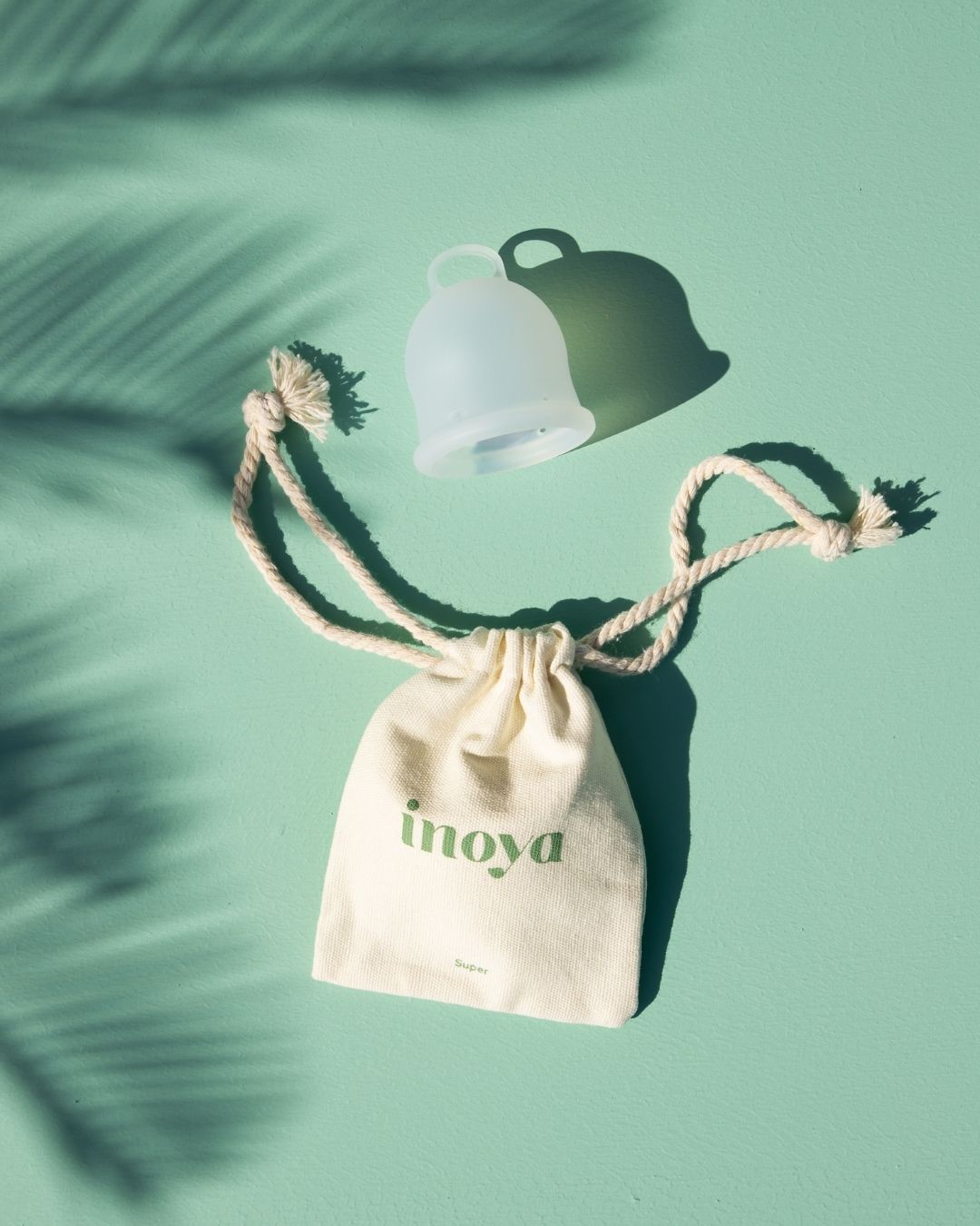 A reusable menstrual cup sits above a small cream drawstring pouch with the brand name 'inoya' printed in green, arranged on a mint green background with palm leaf shadows.