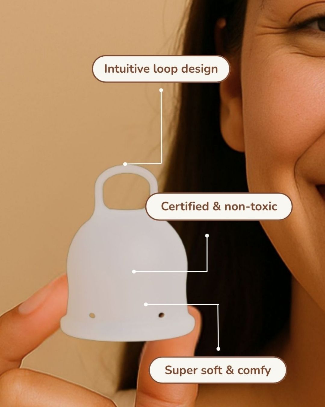 A close-up of a woman holding a translucent white menstrual cup against a warm beige background, with three callout labels pointing to features: 'Intuitive loop design,' 'Certified & non-toxic,' and 'Super soft & comfy.'.