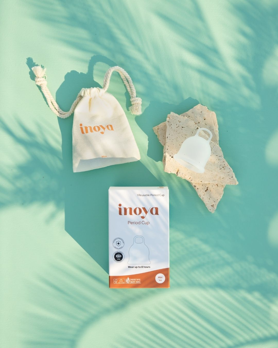 An Inoya Period Cap set arranged on a mint green surface with palm leaf shadows. The set includes a white drawstring pouch with the Inoya logo, a box of Period Cap, and a small white reusable cup resting on textured stones.