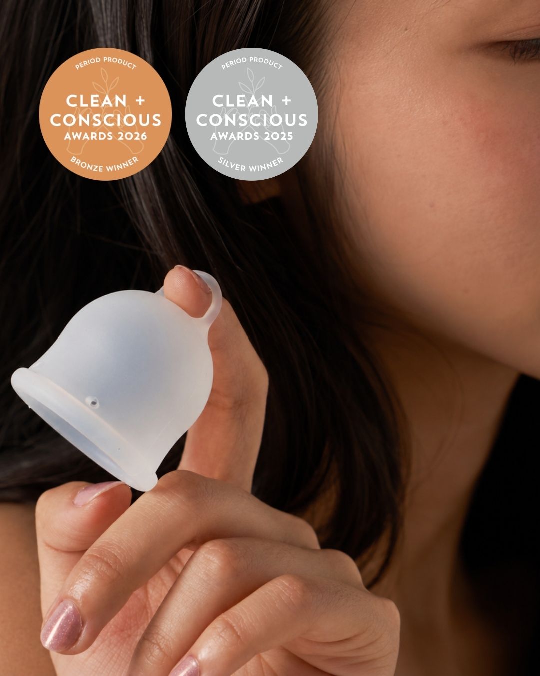 A close-up lifestyle shot of a woman holding a translucent silicone menstrual cup. Two award badges for 'Clean + Conscious Awards' are displayed in the upper left corner.