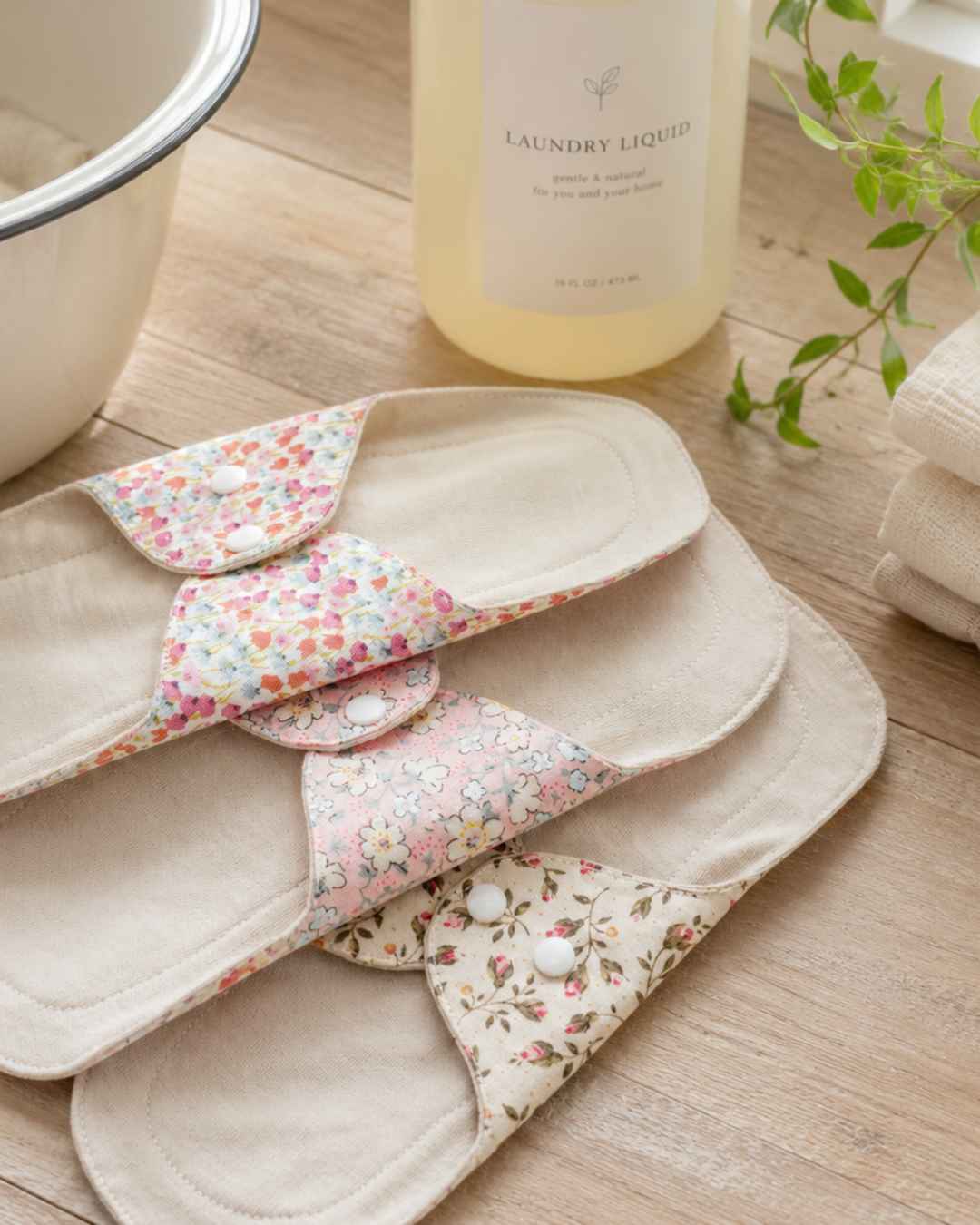three reusable panty liners arranged on a wooden surface next to a laundry liquid bottle and a white enamel bowl.