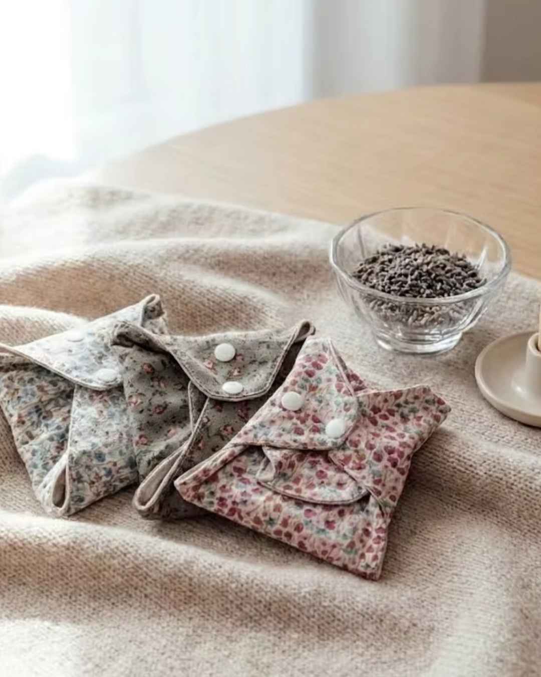 three small reusable pads with floral prints and snap closures resting on a beige textured blanket on a wooden table, with a small glass bowl of dried seeds nearby.