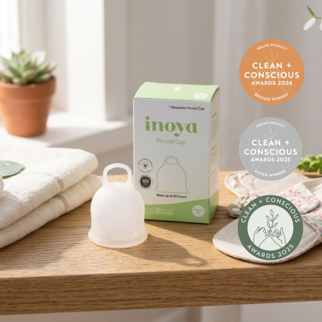 Inoya Period Cup set shown on a wooden table with its product box, a translucent white silicone cup, and award badges. The scene includes folded towels, a small potted plant, and a cloth pad in a bright indoor setting.