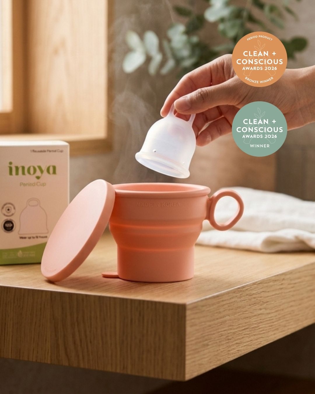 A silicone menstrual cup is shown being held above a matching coral pink collapsible cup on a wooden table, with the product box visible at left and a hand visible at right. Two award badges read 'Clean + Conscious Awards 2024' and 'Clean + Conscious Awards 2025 Winner.